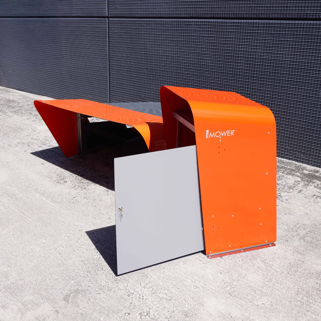 Außeninstallation des technischen Schranks für Mähroboter neben der Garagenstruktur. Das auffällige orangene Design schützt Werkzeuge und Ersatzteile mit einem Schloss an der Front.